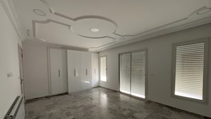 A spacious room with a marble floor, white walls, and a ceiling with decorative molding. There are sliding doors and a window with blinds. - Villa The Crown  (Ariana)