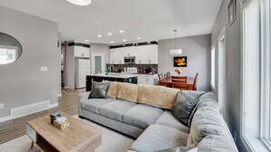 A modern living room with a grey sectional sofa, a wooden coffee table, and a kitchen in the background. - Luxury in Laurel-8 guests+1 Baby,4 beds,2.5 bath,Dbl Grg,discount w direct book. (Edmonton)