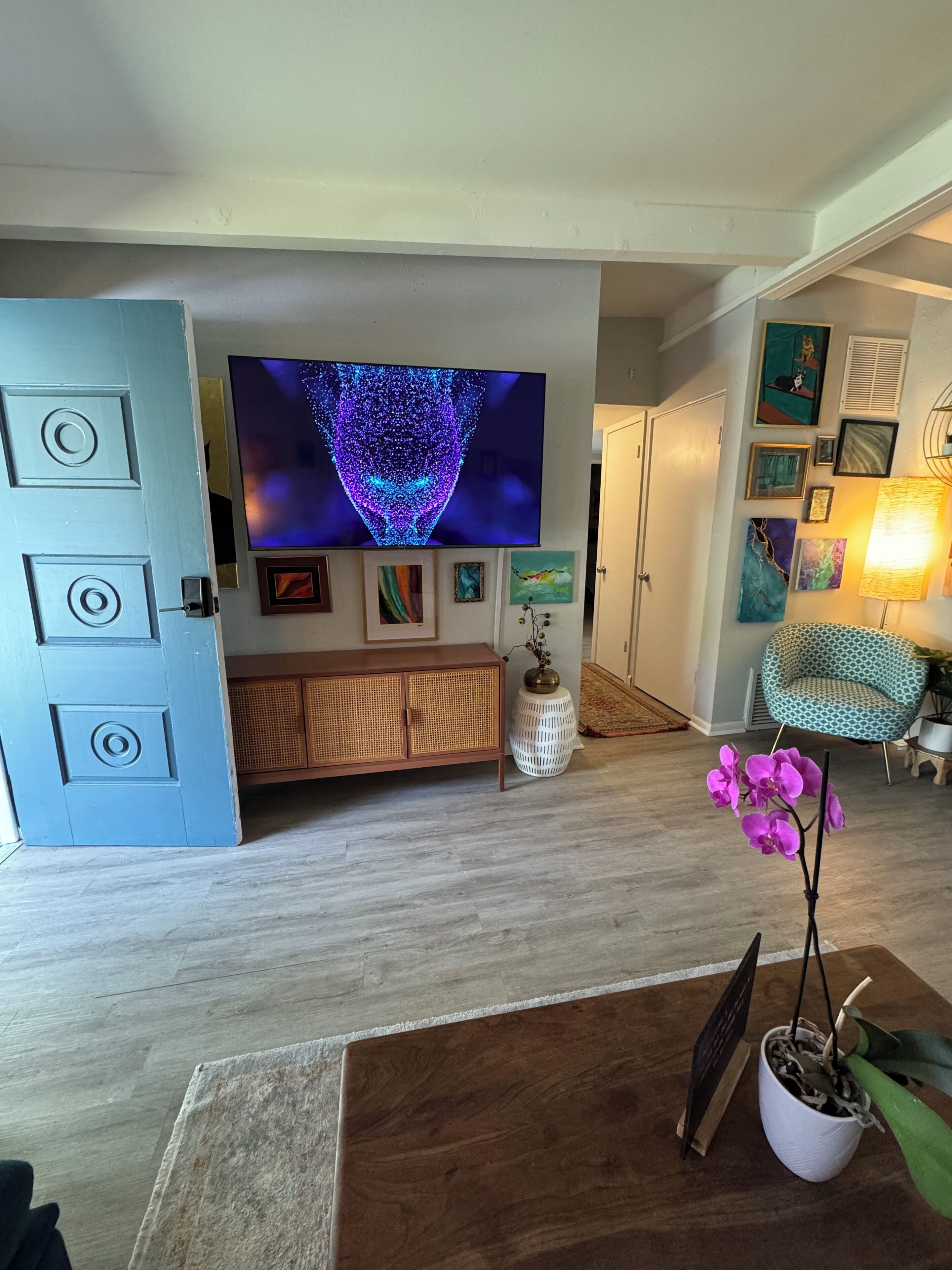Ein Wohnzimmer mit Flachbildfernseher, einer Rattan-Kommode, einer weißen Vase mit Pflanze, einem Holztisch und einem gemusterten Sessel.