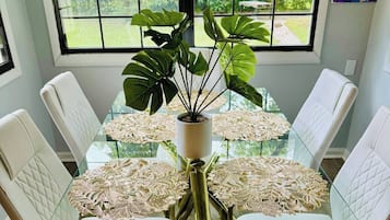 A dining area with a glass table surrounded by white cushioned chairs, a central potted plant, and intricate doilies on the table.