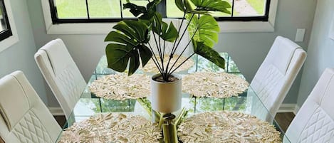Un comedor con una mesa de vidrio rodeada de sillas blancas con cojines, una planta en maceta en el centro y delicados manteles individuales sobre la mesa.