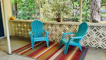 A covered outdoor seating area with two turquoise chairs, a colorful striped rug, and a view of greenery and trees.