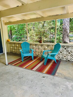A covered outdoor seating area with two turquoise chairs, a colorful striped rug, and a view of greenery and trees. - Nina Juniper Art House Exhibit (Gainesville)