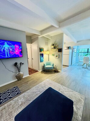 A modern living room with a flat-screen TV, a blue armchair, and a dining area with a table and chairs. - Nina Juniper Art House Exhibit (Gainesville)