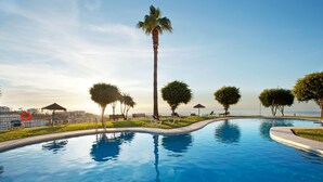 Ein Pool mit geschwungener Kante, umgeben von Bäumen und Bänken, mit Blick auf das Meer im Hintergrund.