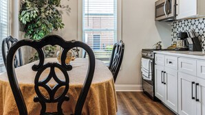 Un coin repas avec une table recouverte d’une nappe beige, entourée de chaises à dossiers décorés. On aperçoit une plante en pot à l’arrière-plan.