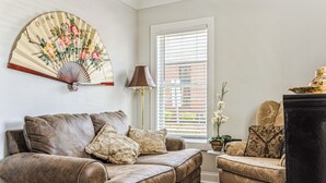 Un salon avec un canapé en cuir marron, un fauteuil assorti, un ventilateur traditionnel orné de motifs floraux, une lampe sur pied et une fenêtre avec des stores.