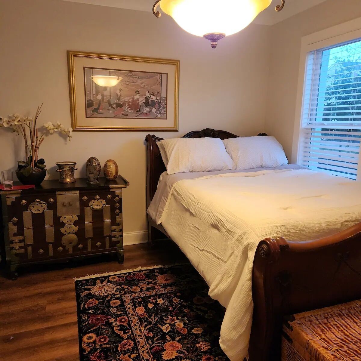 A bedroom with a wooden bed, a nightstand, a dresser, a framed picture, and a window with blinds.
