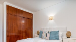 Un dormitorio con un armario de madera, una cama con varios almohadones, una lámpara de noche y una lámpara empotrada en la pared.