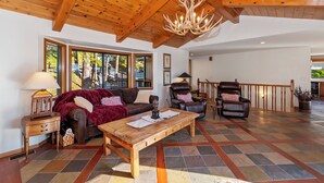 Un salón amplio con techo de madera, una lámpara de araña grande, una chimenea y vistas a árboles a través de ventanas grandes.