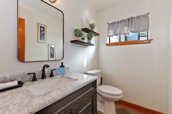 A bathroom with a marble sink, black faucets, a white toilet, a mirror, and a window with a curtain.