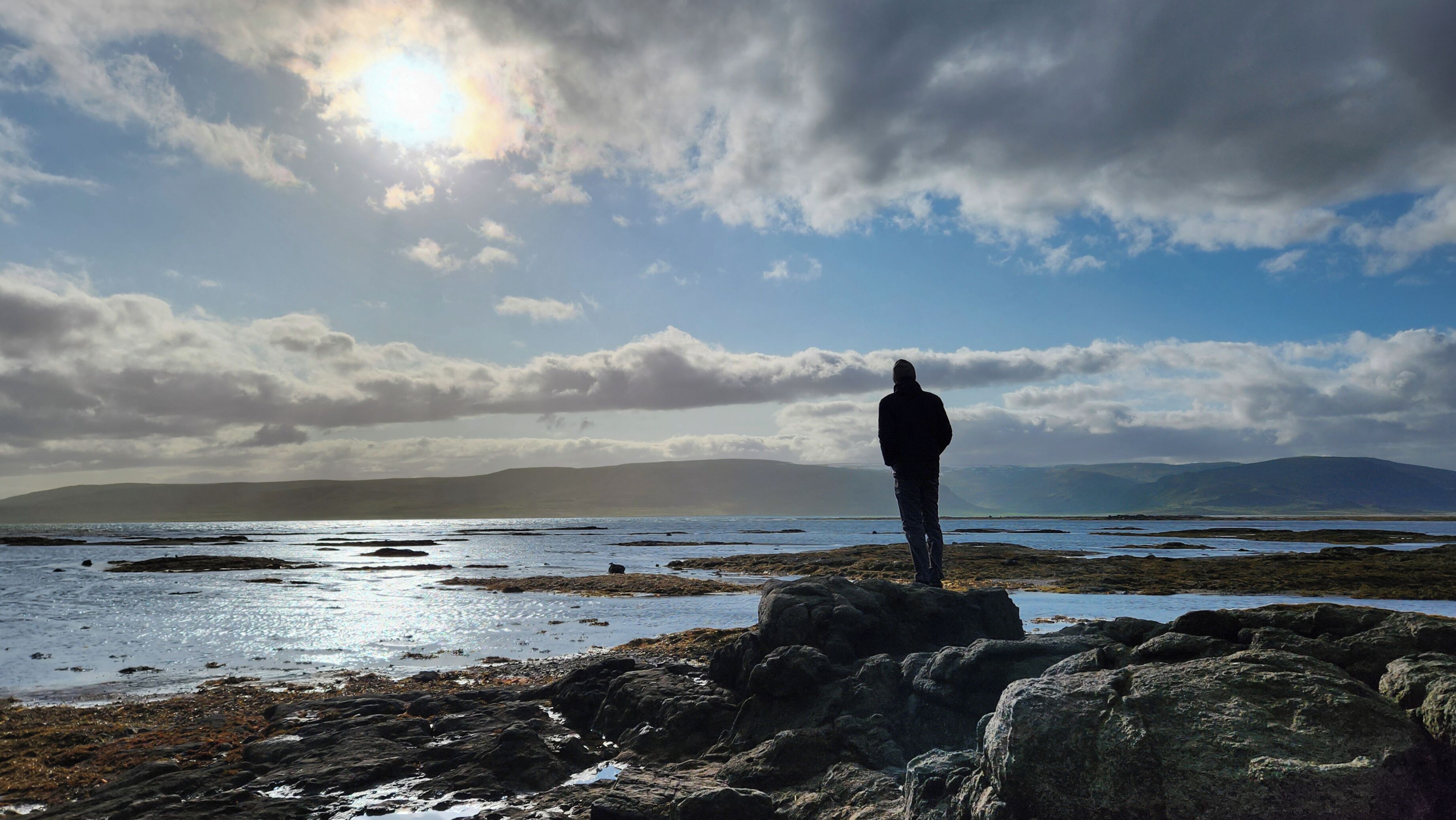A person standing on rocky terrain with a view of a body of water and distant mountains.