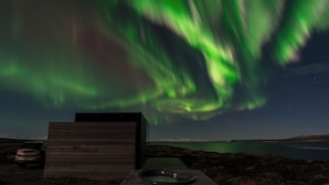 A wooden cabin with a hot tub and a car parked outside, overlooking a body of water with the aurora borealis in the background. - Mystic Light Lodge (Budardalur)
