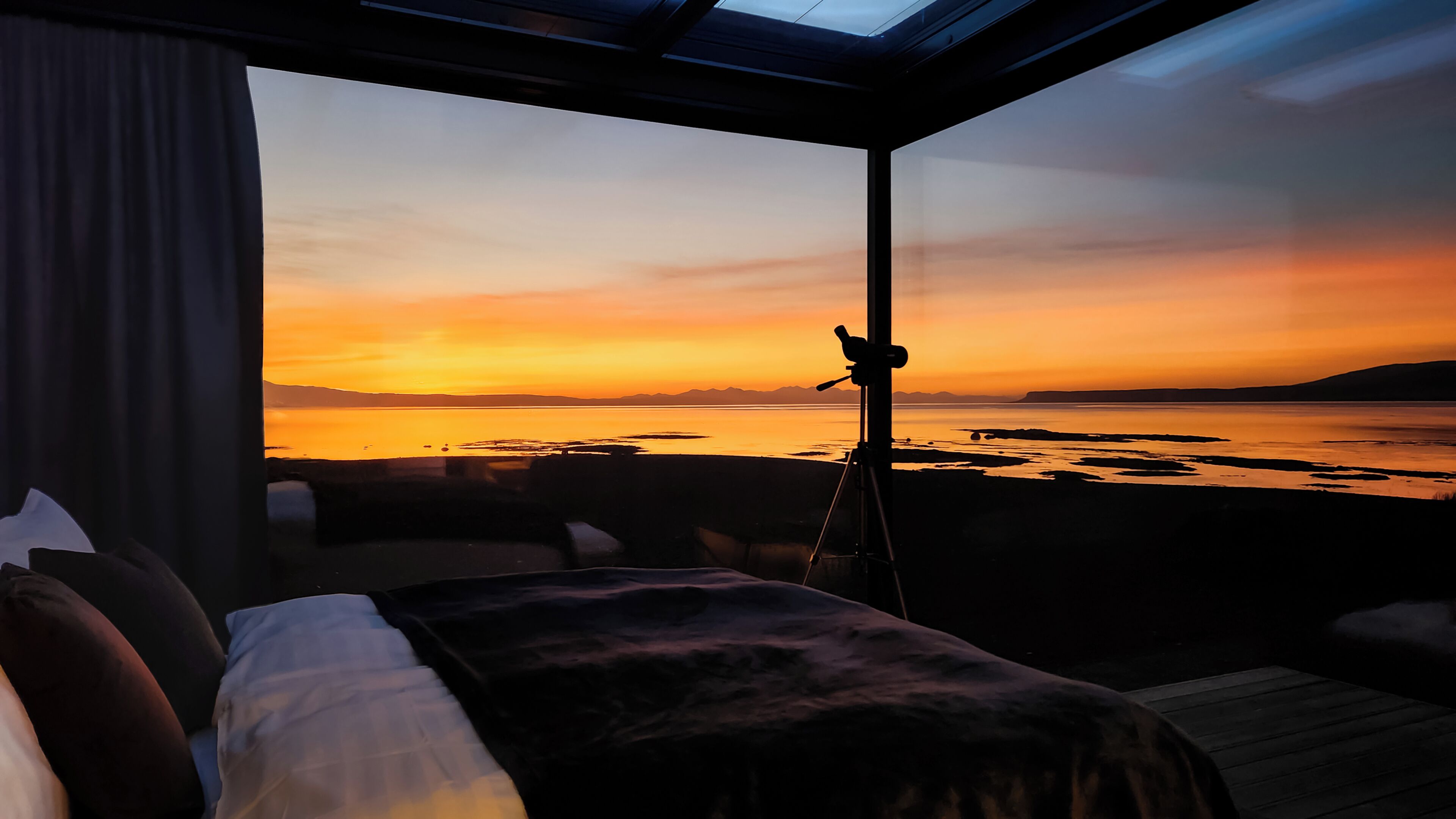 A room with a bed, a window, and a view of a sunset over water.