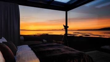 A room with a bed, a window, and a view of a sunset over water.