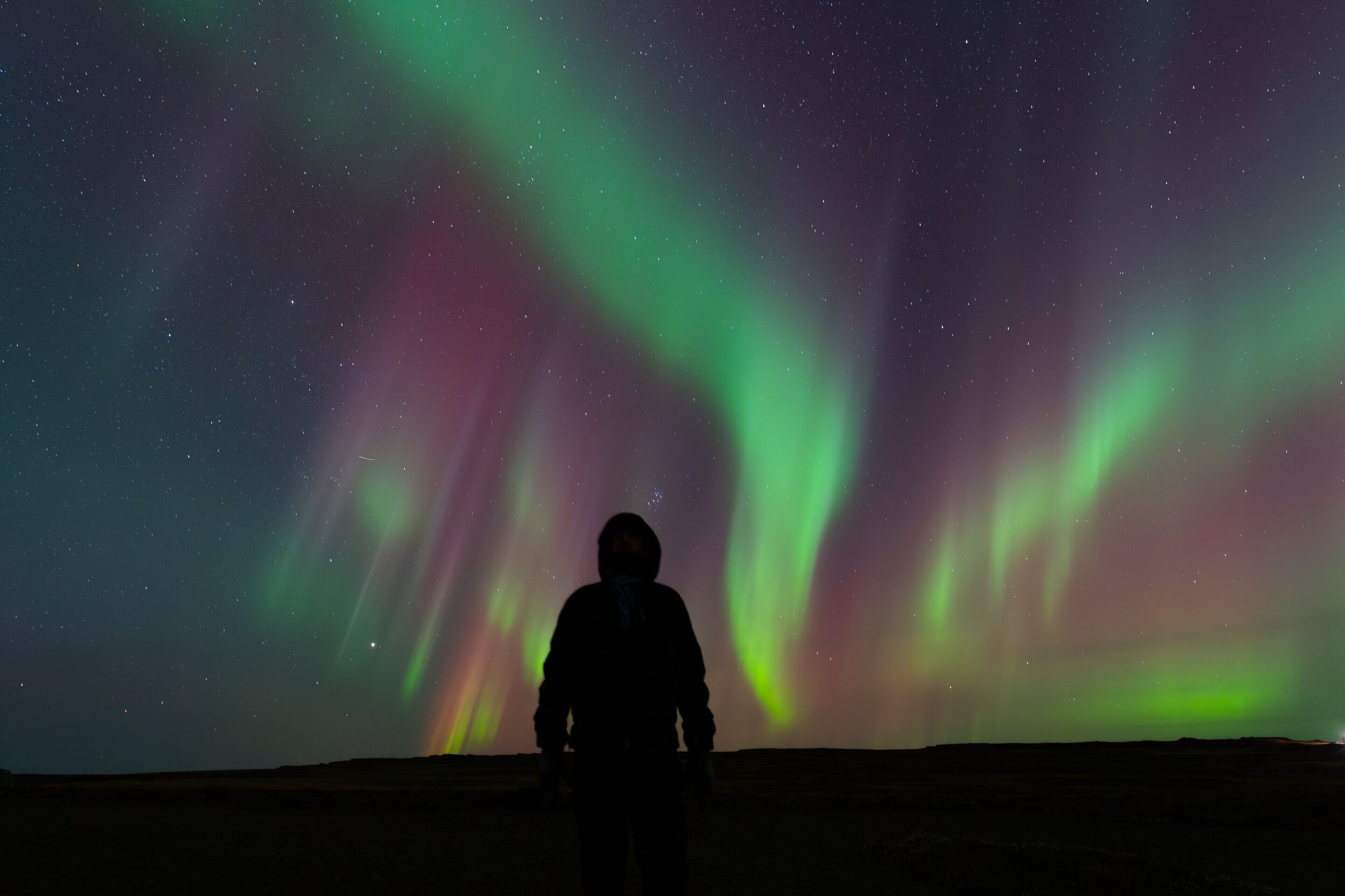 Una persona de pie en un paisaje extenso y abierto bajo un cielo estrellado con una aurora boreal vibrante.
