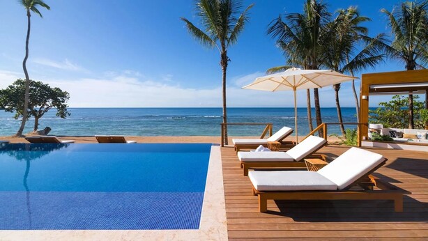 Zona junto a la piscina con sillones de descanso, terraza de madera y vista al océano.
