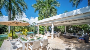 Un patio al aire libre con una mesa de comedor, sillas y una zona de estar cubierta con sofás y una piscina al fondo.