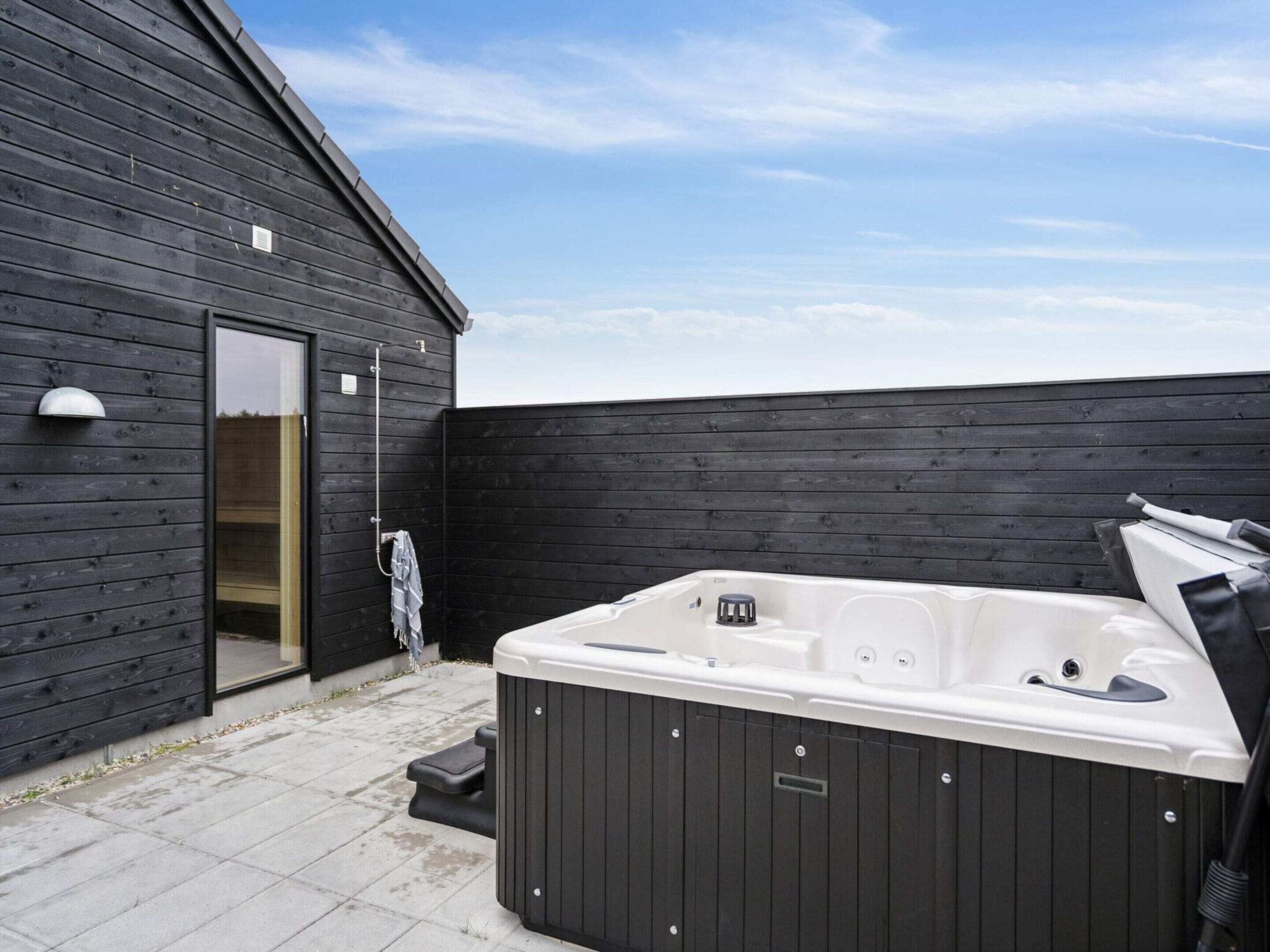 Et moderne udeområde med jacuzzi, træterrasse, en sort trævæg og en klar himmel.