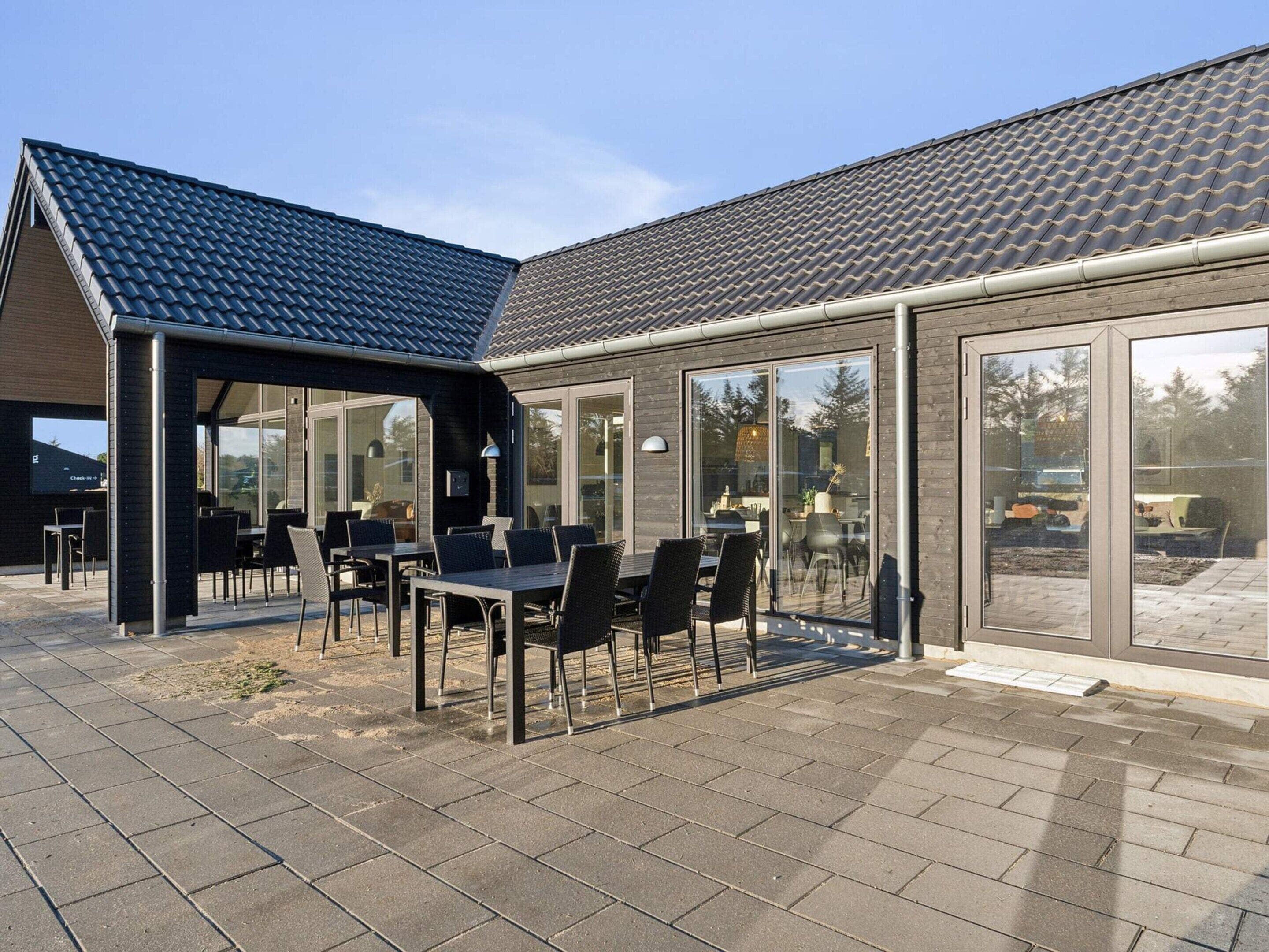 En moderne bygning med tegltag, store glasruder og en patio med udendørs siddepladser.