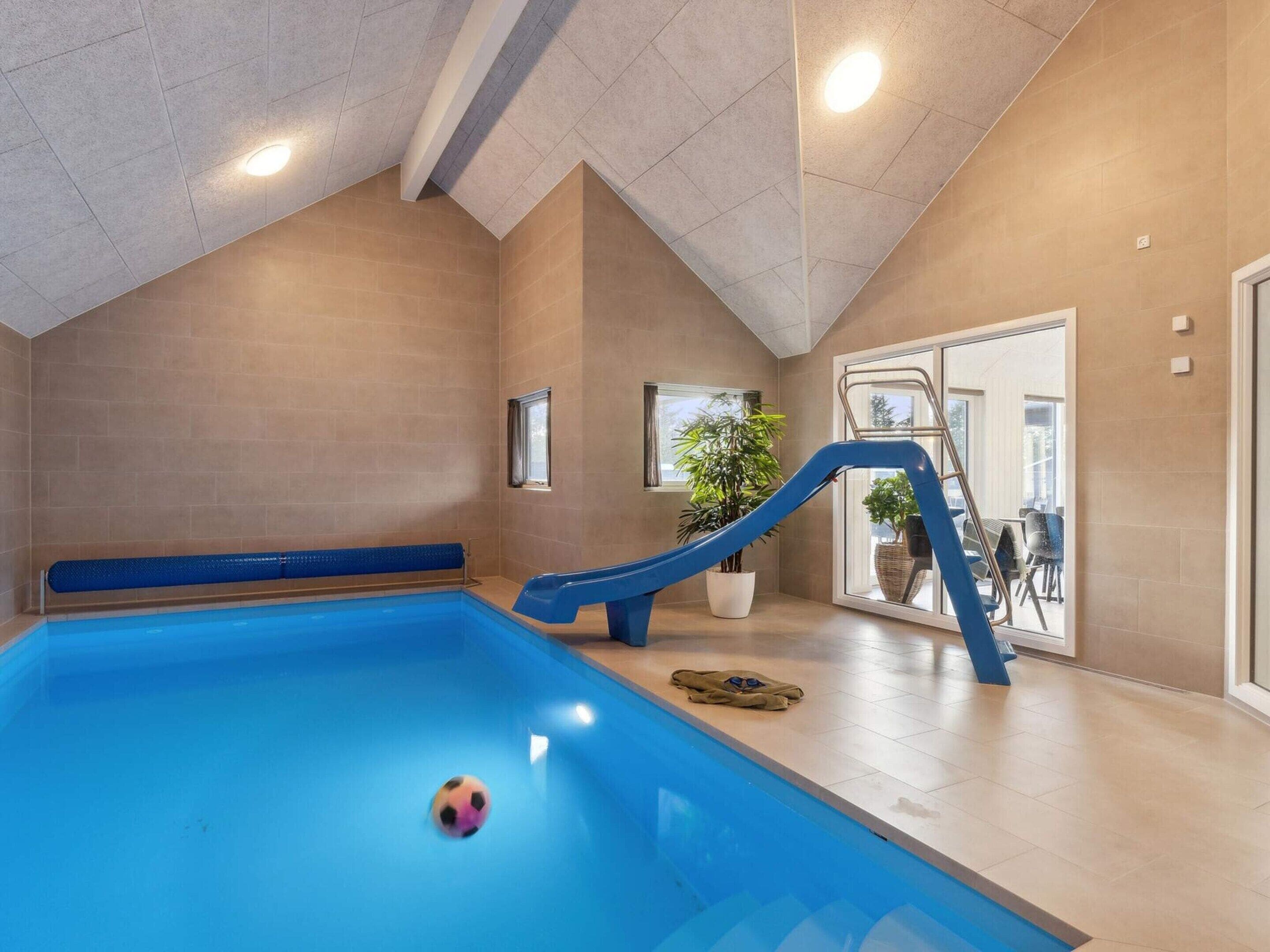 En indendørs swimmingpool med blå rutsjebane, et lille vindue og en potteplante.