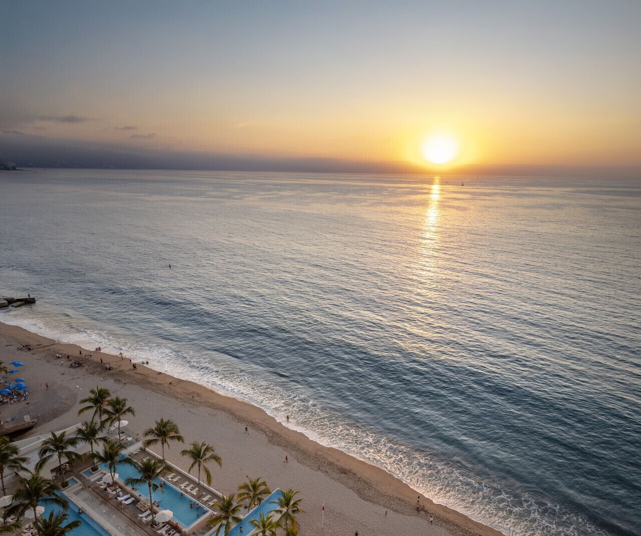 A beach with a pool area, palm trees, and a sunset over the ocean.