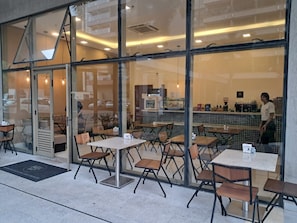 Ein gemütliches Café mit Tischen, Stühlen und einer Theke, hinter der eine Person steht, sowie einer gefliesten Rückwand.