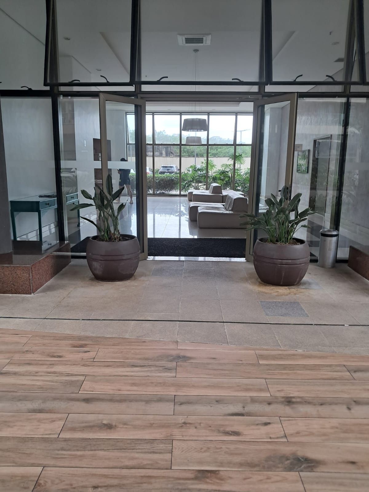 A modern lobby with a glass door leading to a seating area, potted plants, and a tiled floor.
