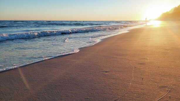 A beach with waves gently lapping onto the shore and a person standing in the distance.