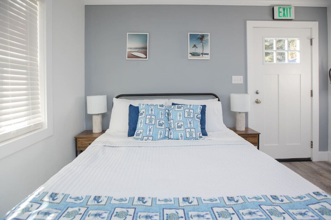 Quarto com cama, duas mesinhas de cabeceira, porta branca com placa de saída e duas quadros nas paredes.