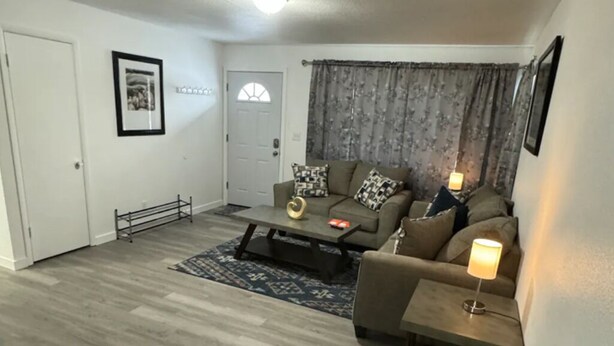 Una sala de estar con un sofá, una mesita de centro y una lámpara. Hay una puerta y una ventana con cortinas.