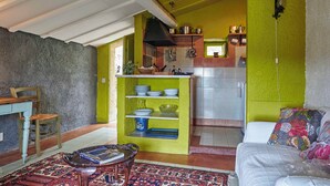 Uma sala de estar acolhedora com um sofá, um tapete estampado e uma cadeira de madeira. Uma cozinha com prateleiras e fogão é visível ao fundo.