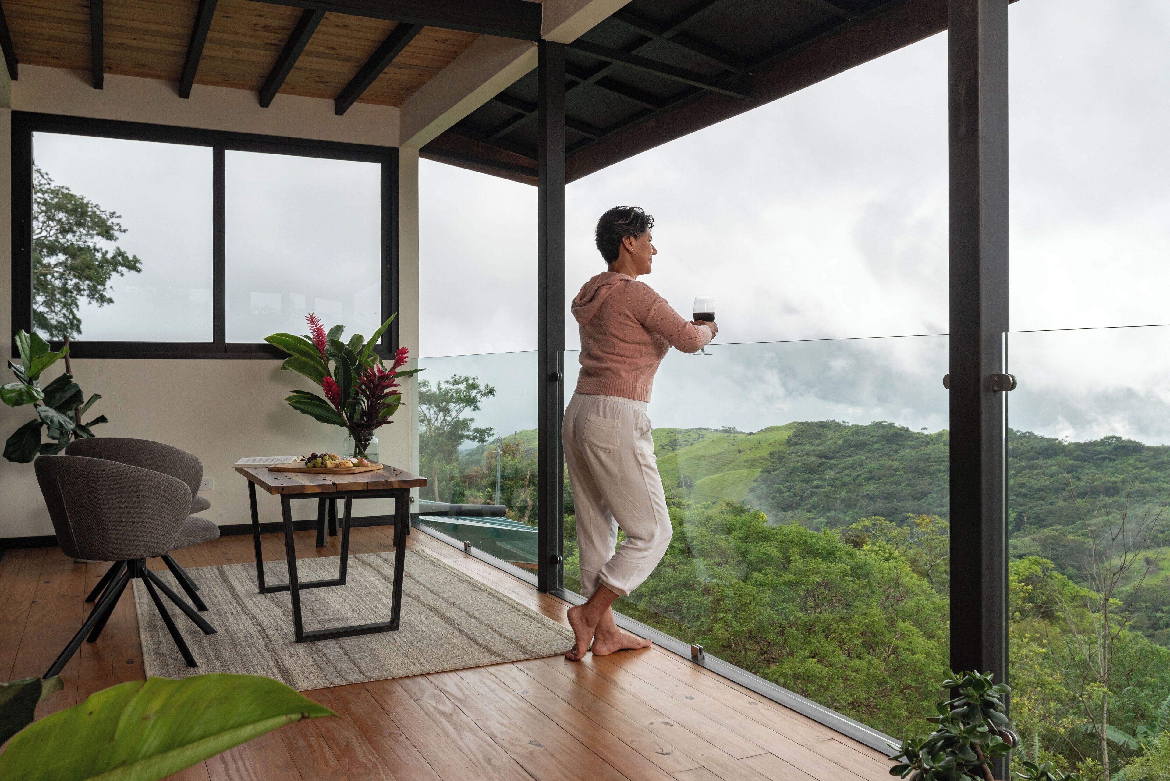 A person standing on a balcony with a glass railing, holding a glass of wine, overlooking a lush green landscape.