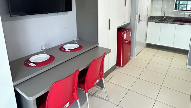 A compact kitchen with a dining area, a flat-screen TV mounted above the counter, and a red refrigerator. - Melhores Flats - Varanda frente ao mar (João Pessoa)