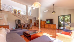 Ein geräumiger Wohnbereich mit einer großen Sofagarnitur, einem Esstisch mit Stühlen, einer Küchenzeile mit Schränken und einem Fernseher, sowie einer Treppe mit dekorativer Geländer.
