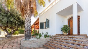 Ein weißes Gebäude mit einer Holztreppe, die zu einer Tür führt, umgeben von einem Garten mit einer Palme und Topfpflanzen.
