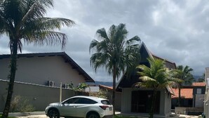 A paved pathway leading to a building with a red-tiled roof, surrounded by palm trees and a white car parked nearby. - Happy Corner Cottage  (Chácaras Mogiano Bertioga)