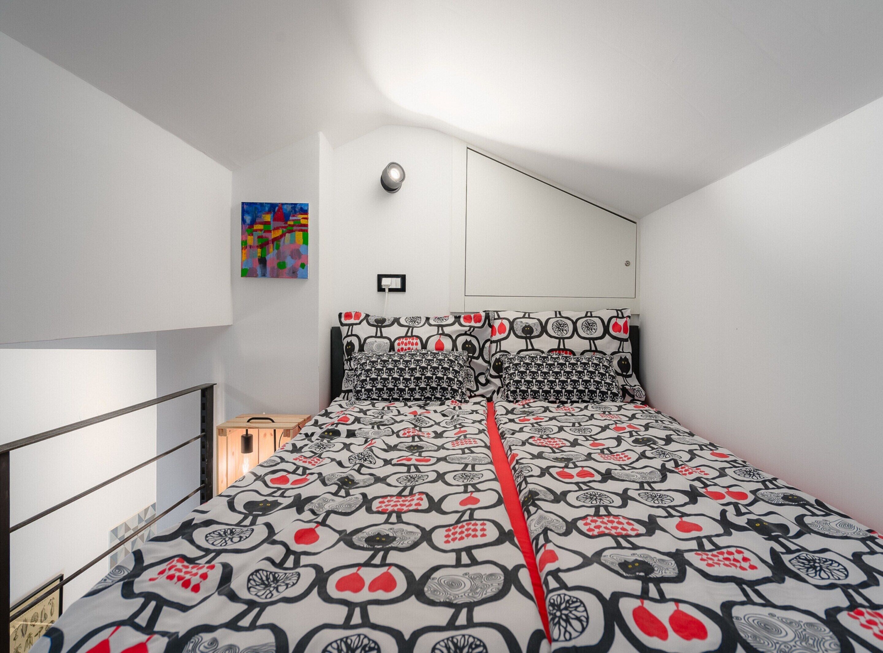 A bedroom with a bed featuring a patterned bedspread, a headboard with a unique design, a small bedside table, and a wall-mounted light fixture.