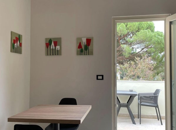 A room with a wooden table and two chairs, a door leading to an outdoor area with a view of trees, and three abstract paintings on the wall.