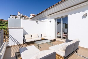 Um pátio moderno ao ar livre, com sofás brancos, uma mesa de centro de vidro e uma porta de correr que dá para um espaço interno.
