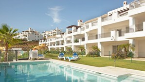 Una piscina con sillones, una estructura de techo de paja y edificios blancos de varios pisos con balcones.