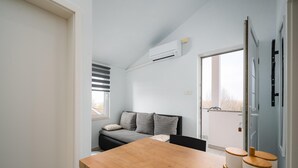 Un espace de vie compact comprenant une table à manger en bois, un canapé avec des coussins, une fenêtre avec des stores et un appareil de climatisation fixé au mur.