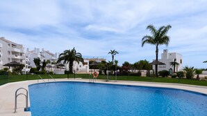 Ein Swimmingpool mit klarem, blauem Wasser, umgeben von einem weißen Gebäude mit Balkonen, Palmen und blauem Himmel.