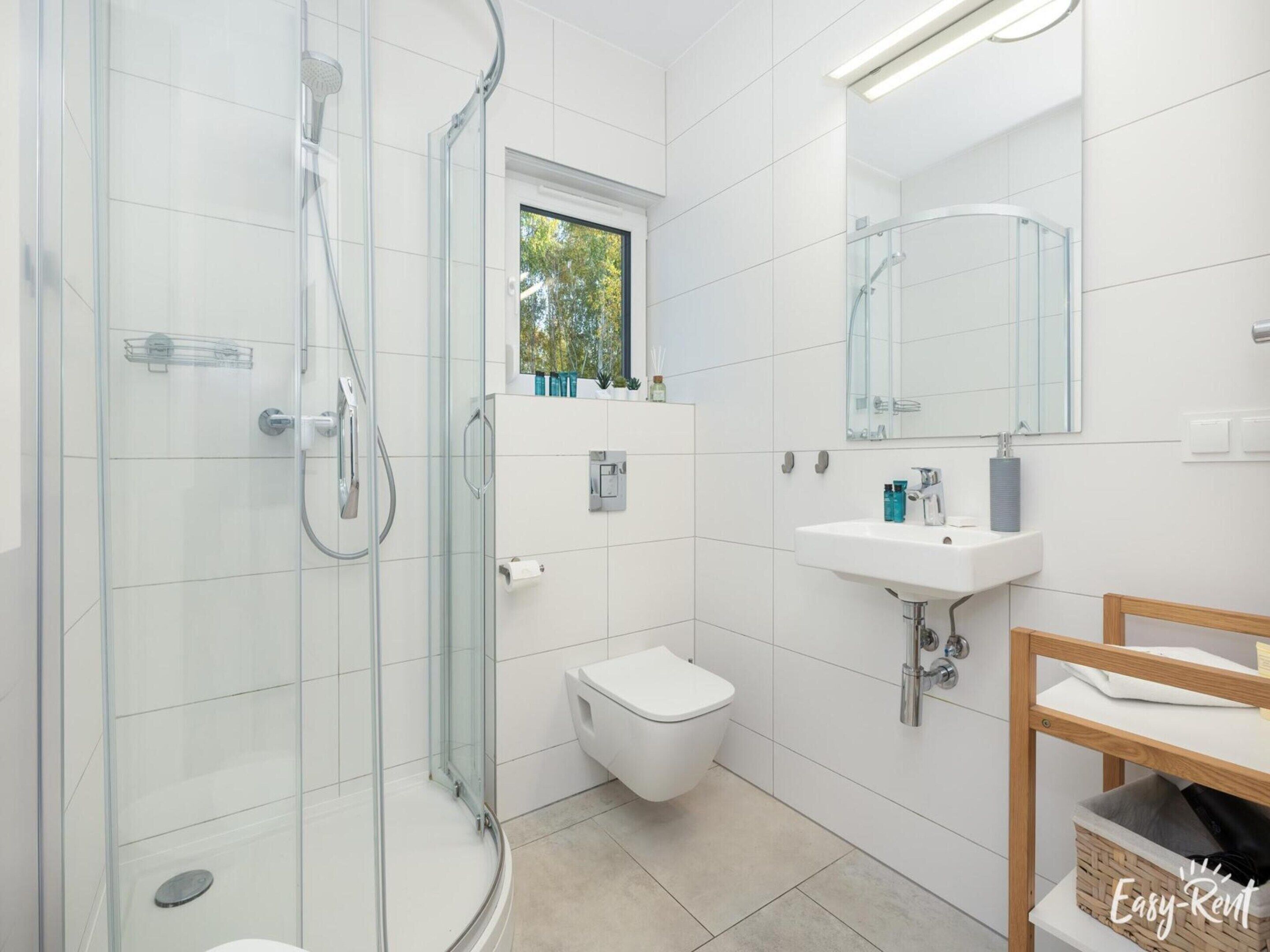 A modern bathroom with a glass-enclosed shower, a toilet, a sink, and a window above the toilet.