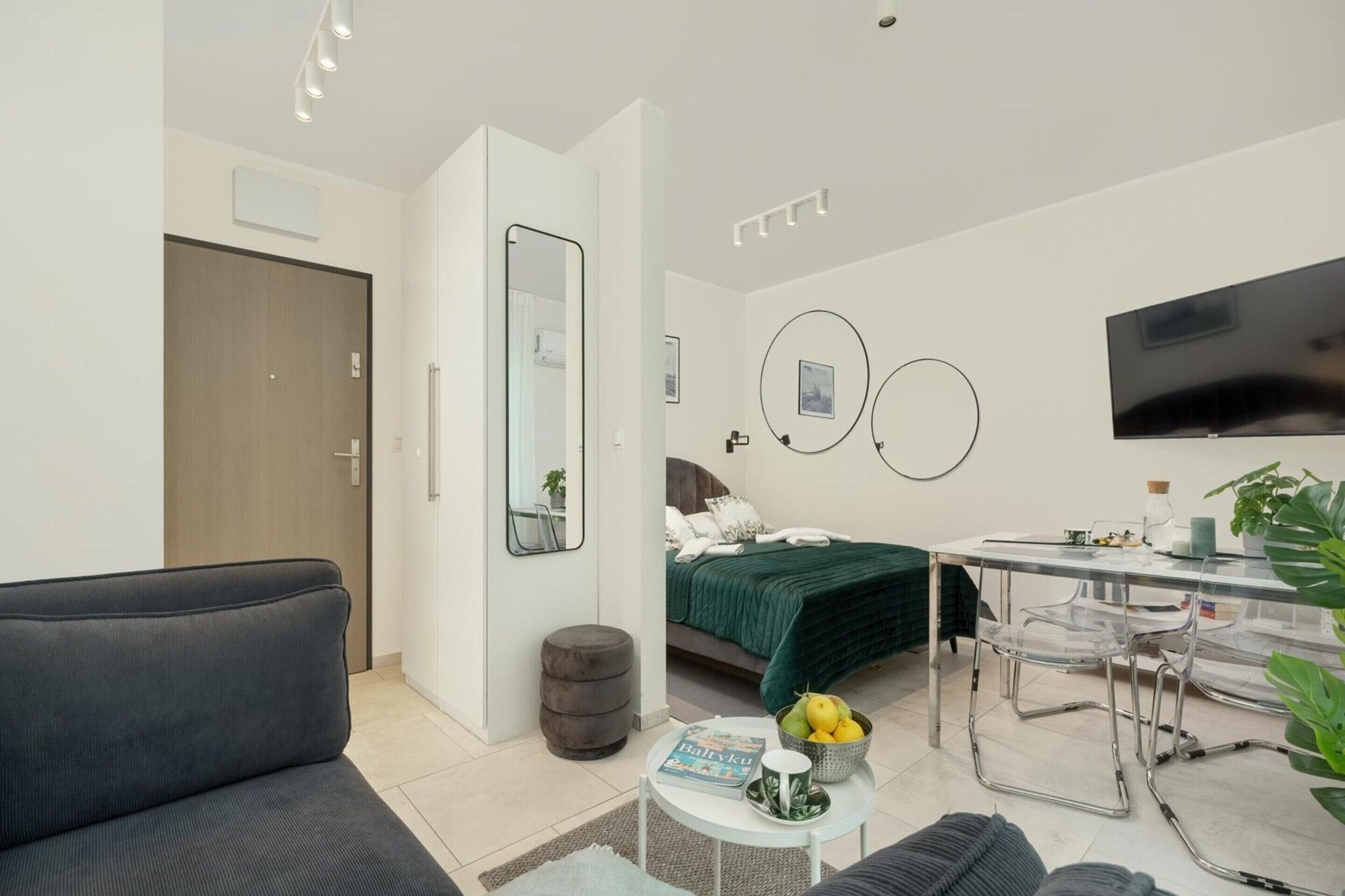 Una stanza moderna e ben illuminata con un divano, un letto con biancheria verde, un tavolo da pranzo con sedie e un televisore montato a parete.