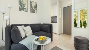 A modern living room with a dark grey sectional sofa, a small white side table with a fruit bowl, and a grey ottoman. There are two framed leaf artworks on the wall, a white floor lamp, and a view of a kitchen and a door leading outside. - Studio in Niechorze Near Baltic Beach (Rewal)