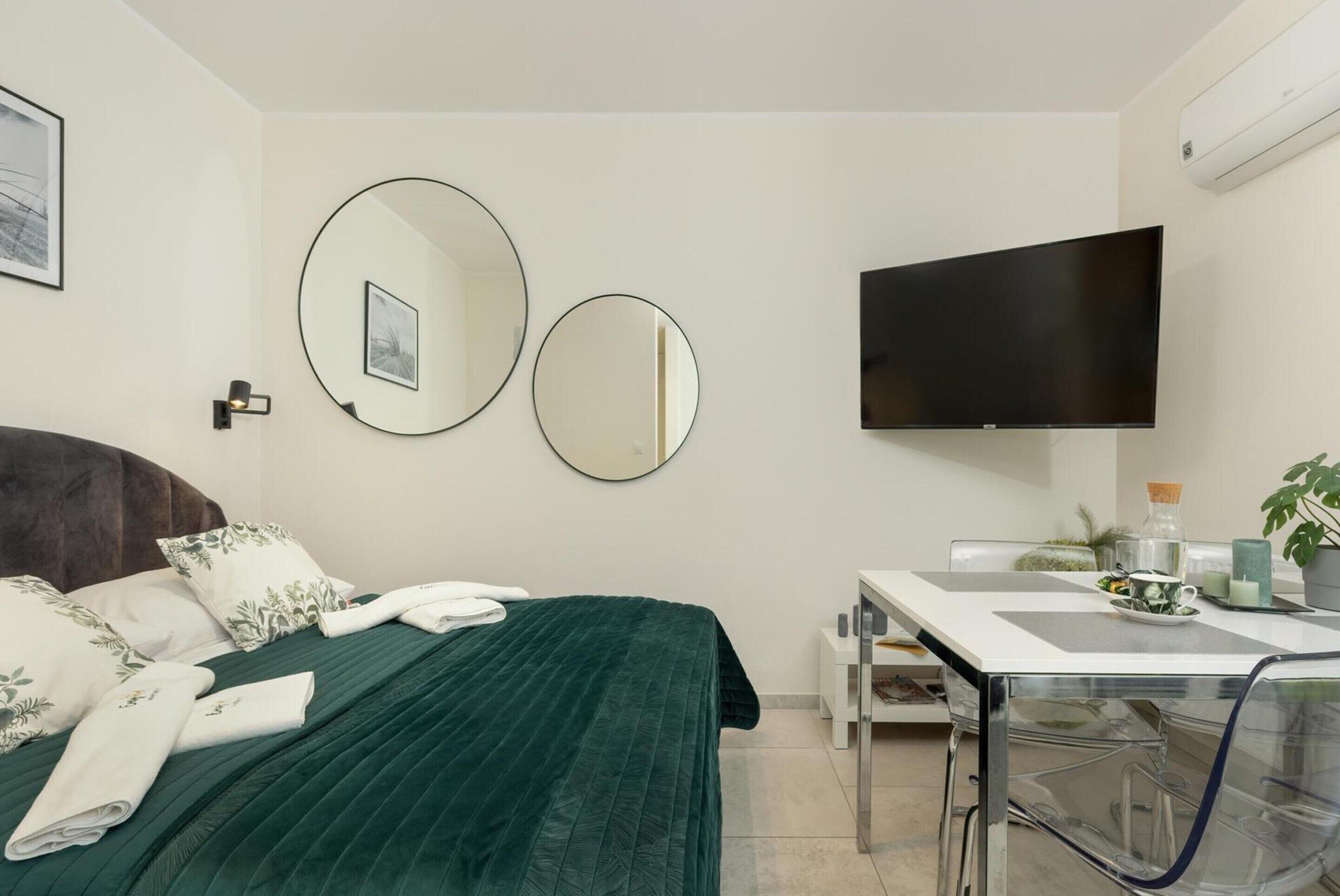 Ein Hotelzimmer mit einem Bett, zwei Wandspiegeln, einem Flachbildfernseher, einem kleinen Tisch mit einer Pflanze und einem Sessel.