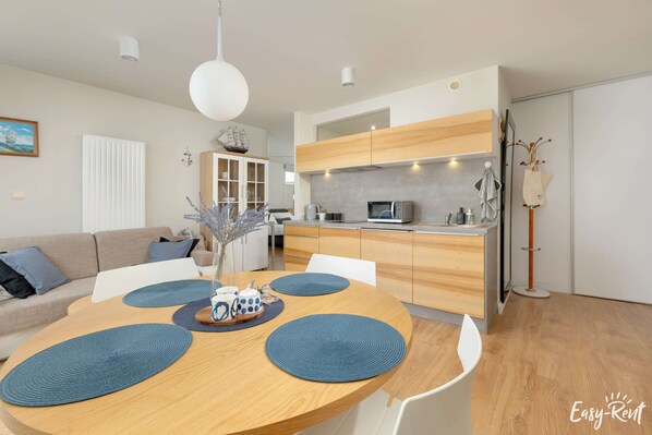 Cocina moderna con armarios de madera, un microondas y un fregadero. Hay una zona de comedor con una mesa redonda y manteles individuales azules.