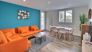 A modern living room with a bright blue accent wall, a large orange sofa, a dining area with a white table and chairs, and a patterned rug. - The Aixoise Escape - Close to the city center (Aix-en-Provence)