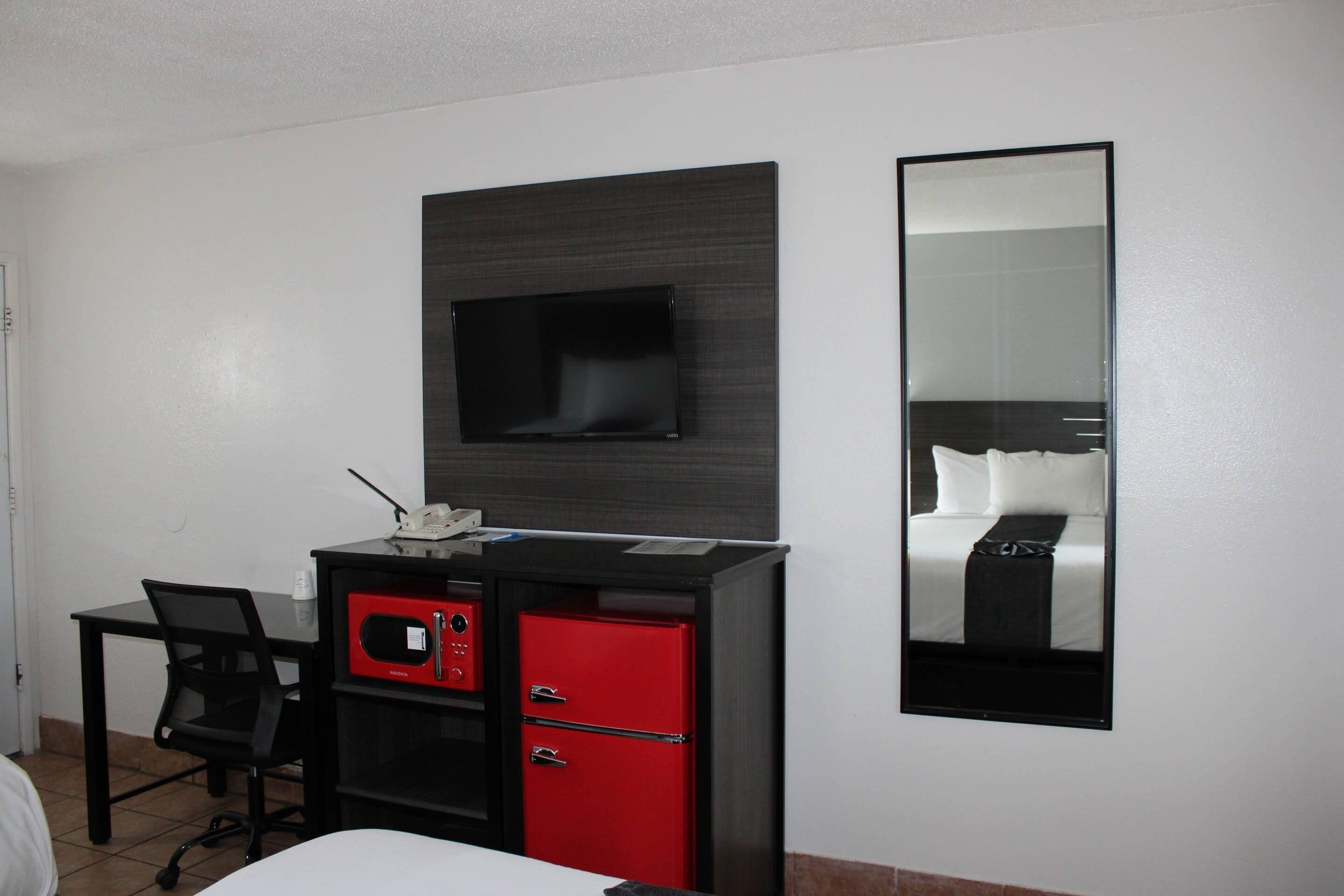Habitación de hotel con cama, escritorio con computadora, archivador rojo, televisor y un espejo grande.